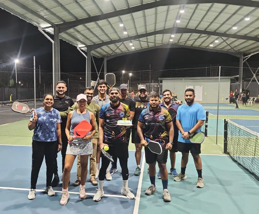 A group of friends celebrating a birthday with a cake and paddles on the floodlit covered courts at night